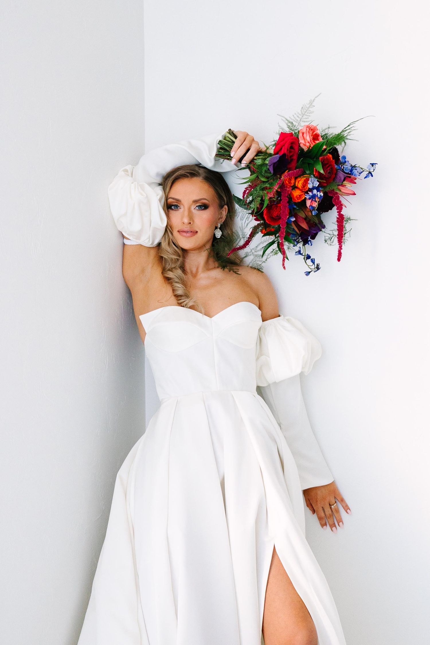 A bride in a classic white wedding dress with dramatic puff sleeves poses with a vibrant red and blue floral bouquet.