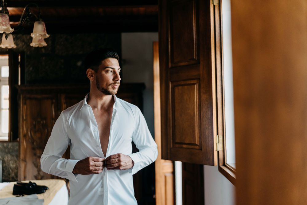 Person in white dress shirt gazes out window in rustic room with natural lighting.