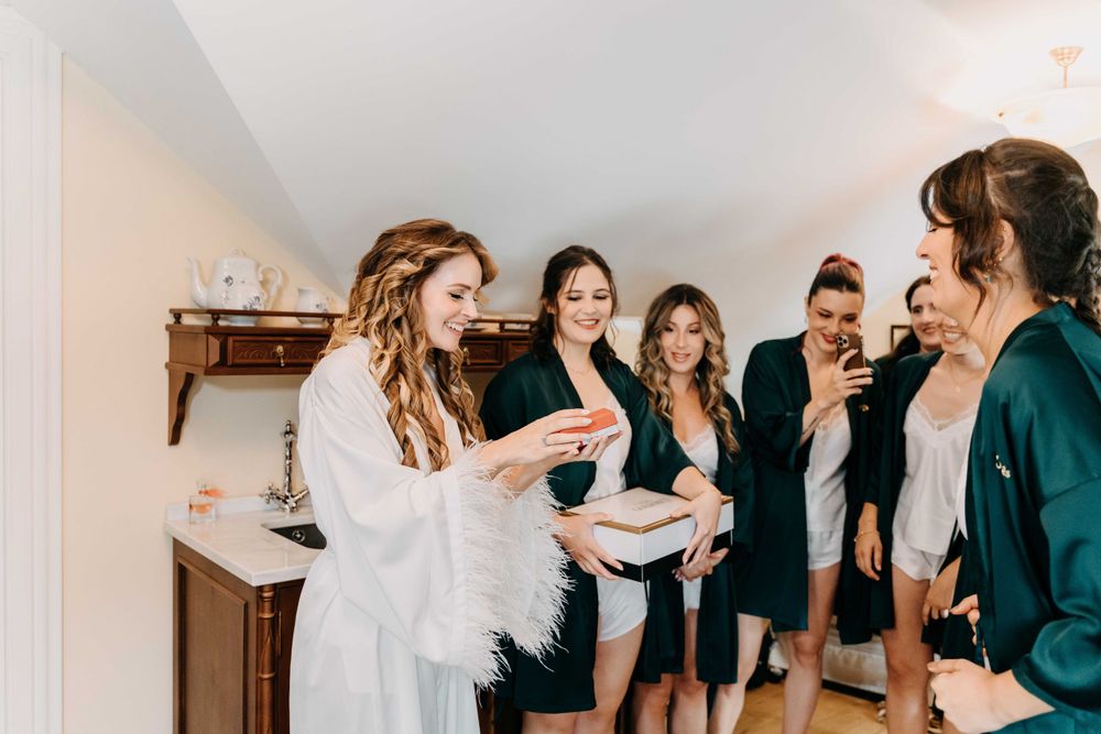 A group of women in robes laugh and smile together while getting ready in a white-walled room.