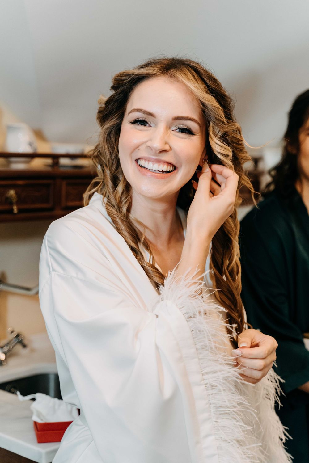 Bride wearing white robe and curled hair smiling brightly while getting ready in bridal preparation room.