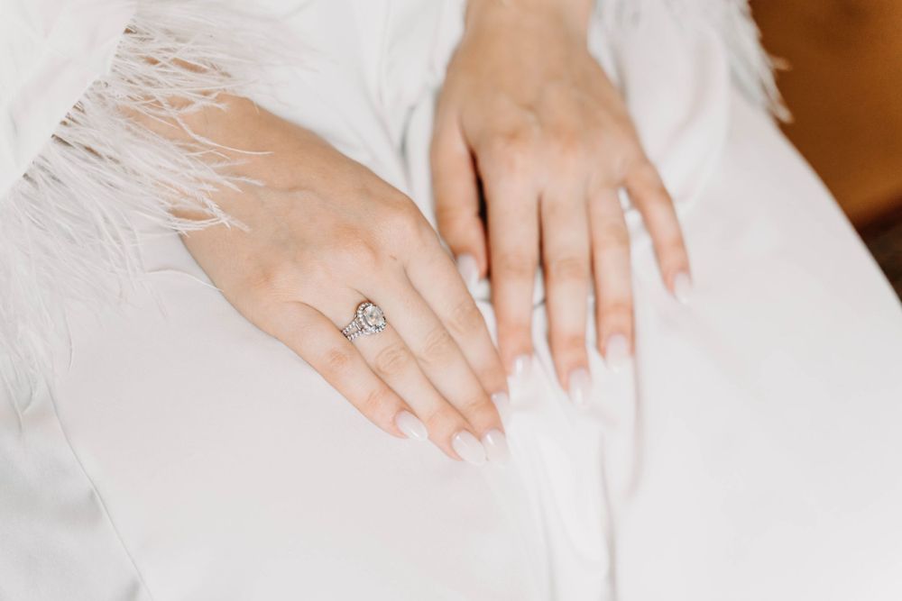 Close up of manicured hands with engagement ring resting on white lace fabric.