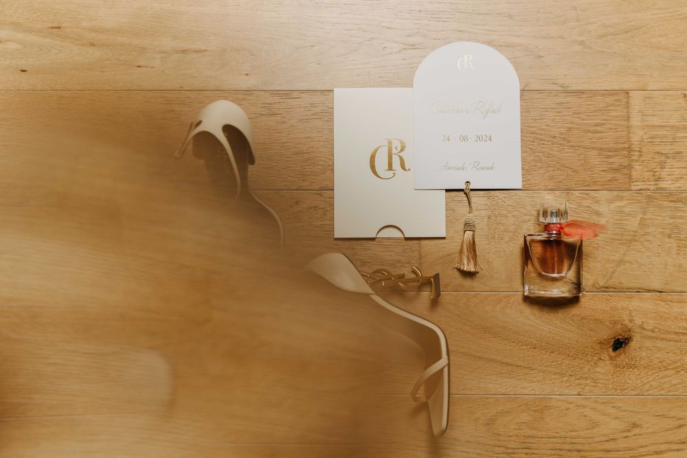 A perfume bottle and wedding invitation resting on a wooden surface with a white hanger in the background.