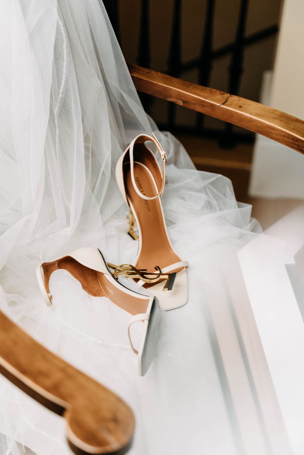White high heel bridal shoes rest on delicate tulle veil fabric draped over a wooden chair.