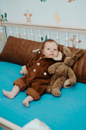 Baby in brown overalls lying on turquoise carpet with plush teddy bear.