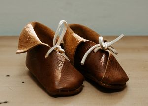 Pair of small brown leather baby shoes with white laces on a neutral background.