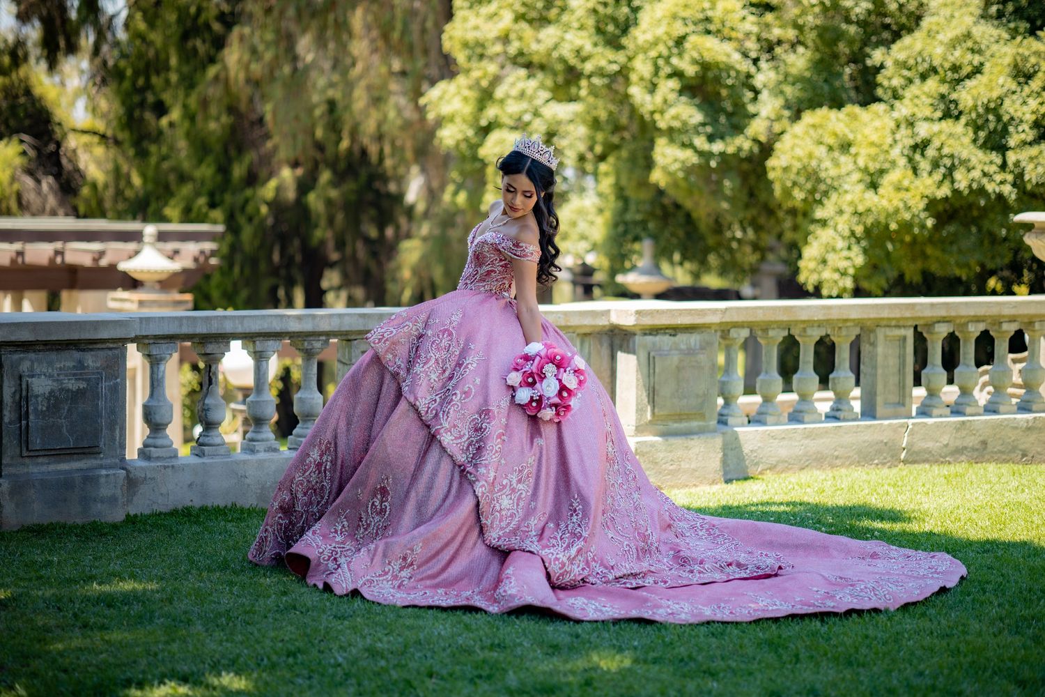 Young woman in elaborate pink quinceañera gown with train poses gracefully on green lawn outdoors.