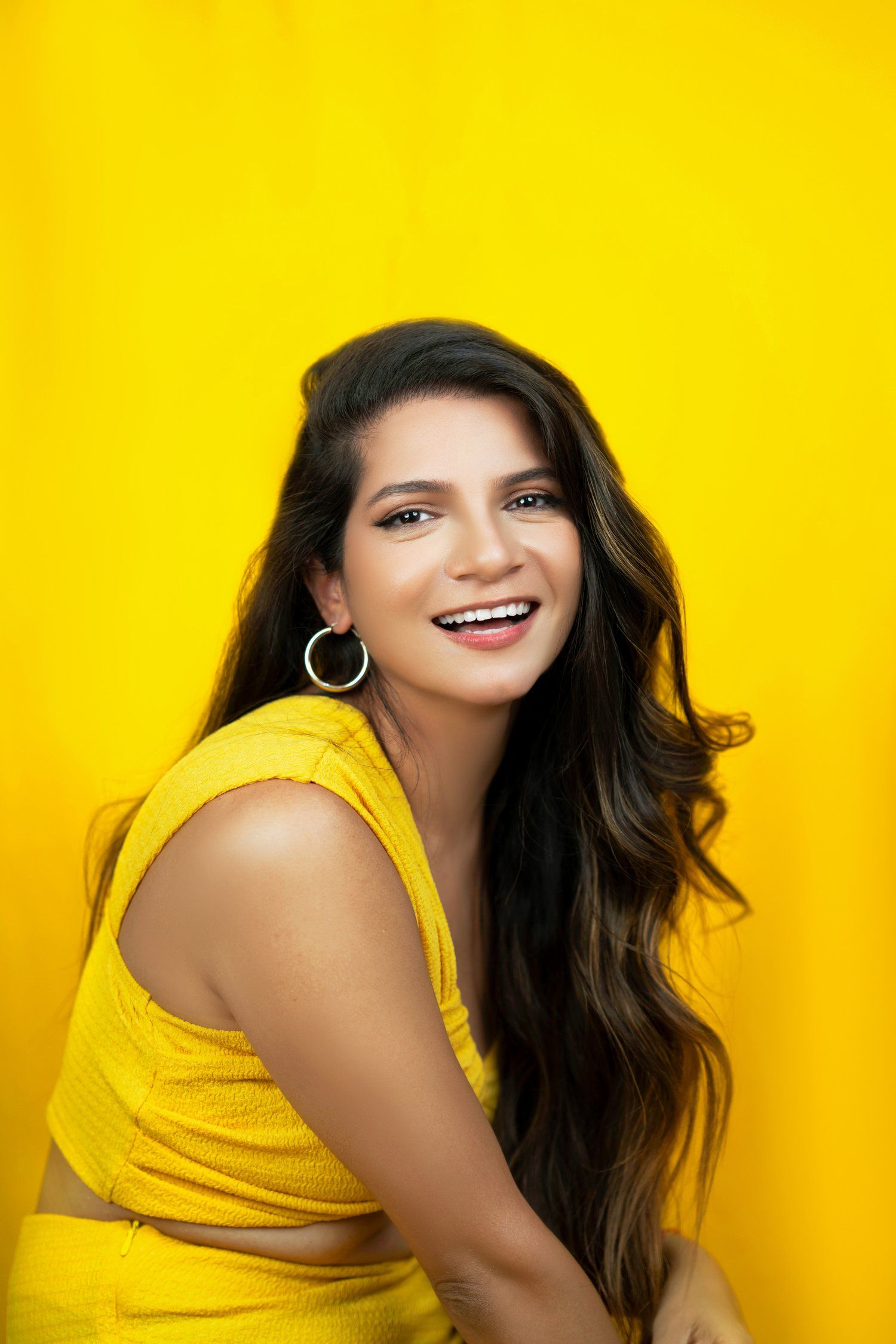 A joyful expression against a bright yellow background, wearing a matching yellow outfit and silver hoop earrings.