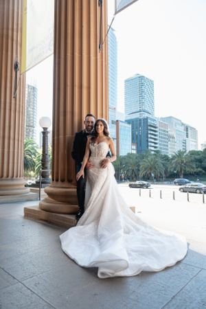 Bride and groom posing by grand columns with city skyline backdrop on their wedding day.