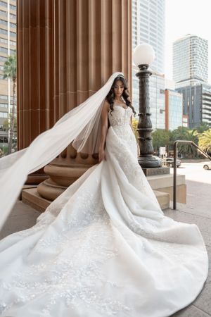 Bride in a floral lace mermaid gown with flowing veil standing beside a classical stone column outdoors.