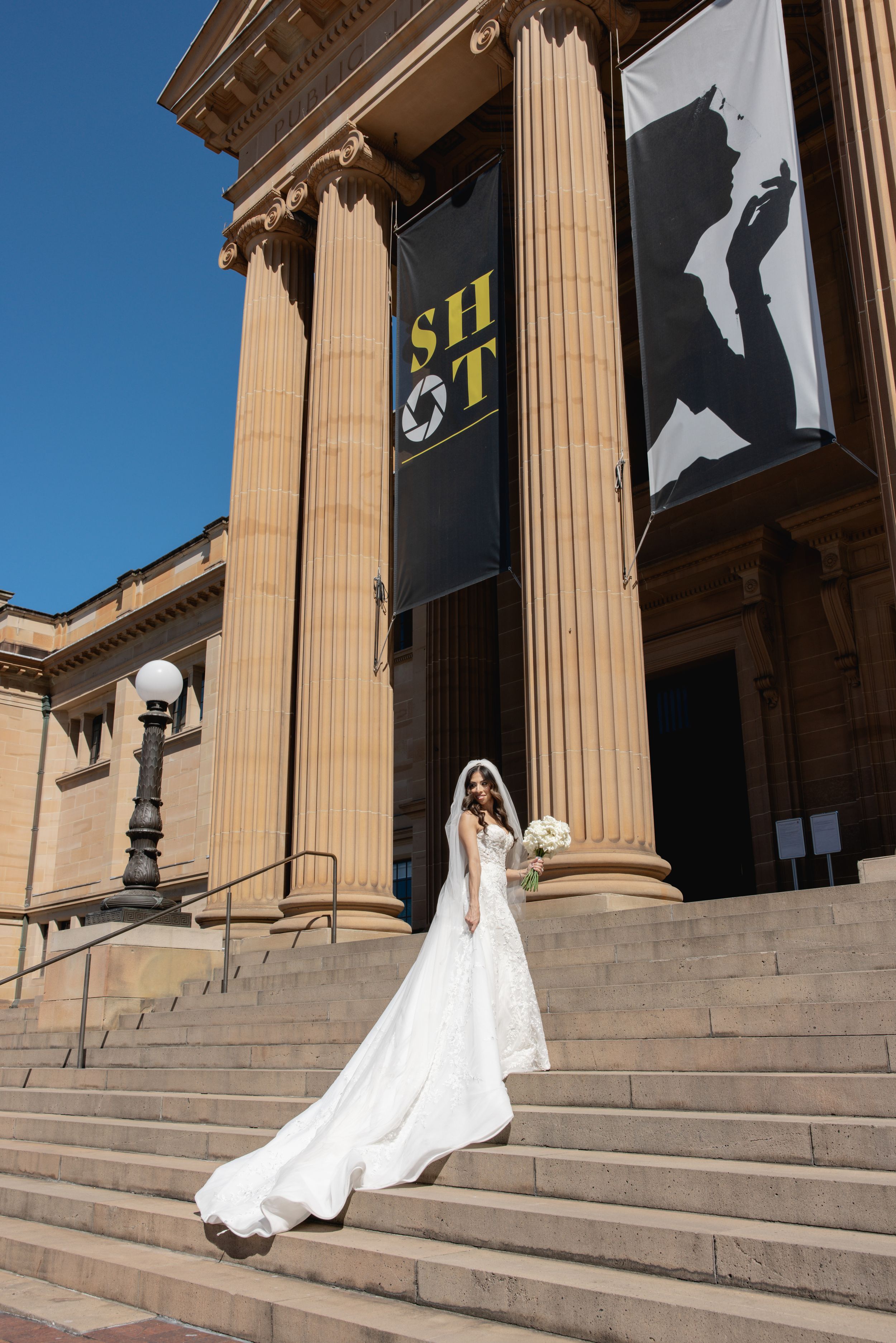 Elegant bride in a flowing white gown and veil standing on steps of a neoclassical building.