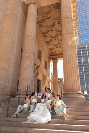 Bride and bridesmaids in sage green dresses sitting on steps of a grand columned building.