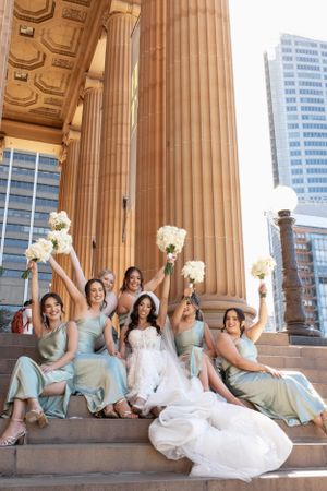Bride and bridesmaids celebrating on steps of a classic building, raising bouquets joyfully.