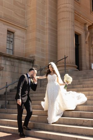 Groom kissing bride's hand on sunlit stone steps of a classic building on their wedding day.