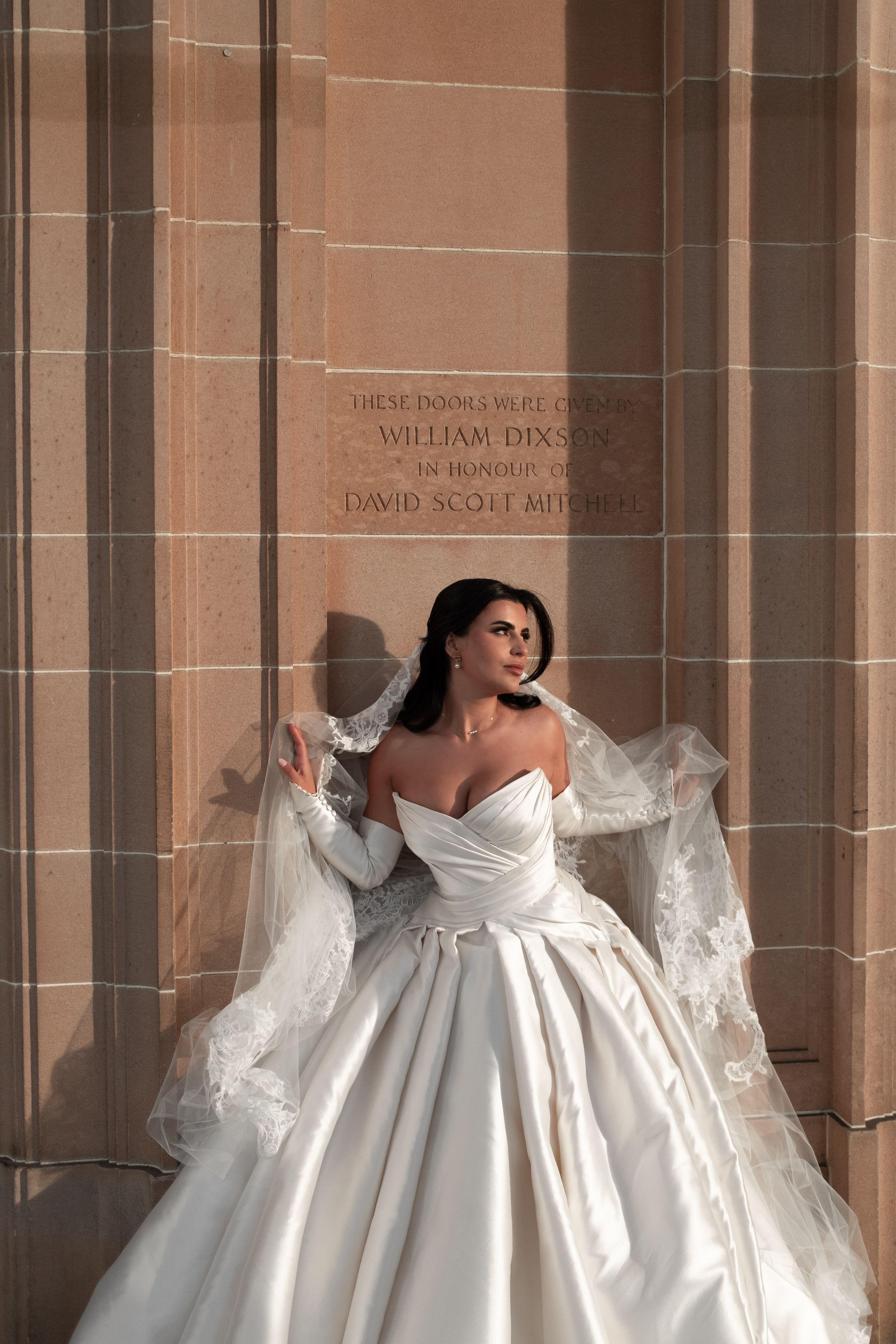 Bride in a sweetheart satin ball gown with lace veil posing against a sunlit sandstone wall.