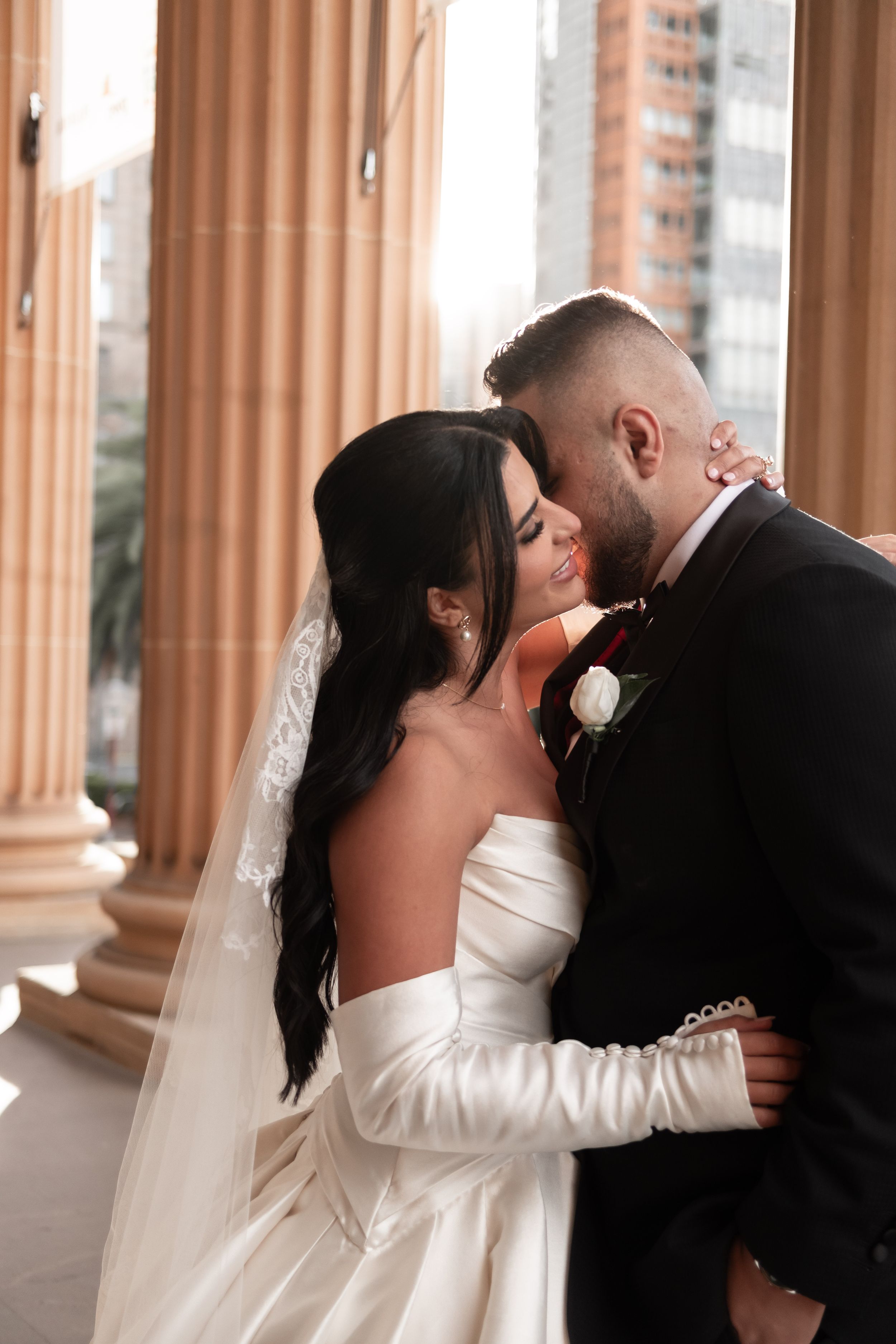Groom tenderly kissing bride's cheek as she smiles beside warm sandstone classical columns.