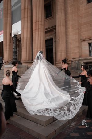 Bridesmaids in black gowns holding the lace-edged cathedral veil of a bride on grand stone steps.