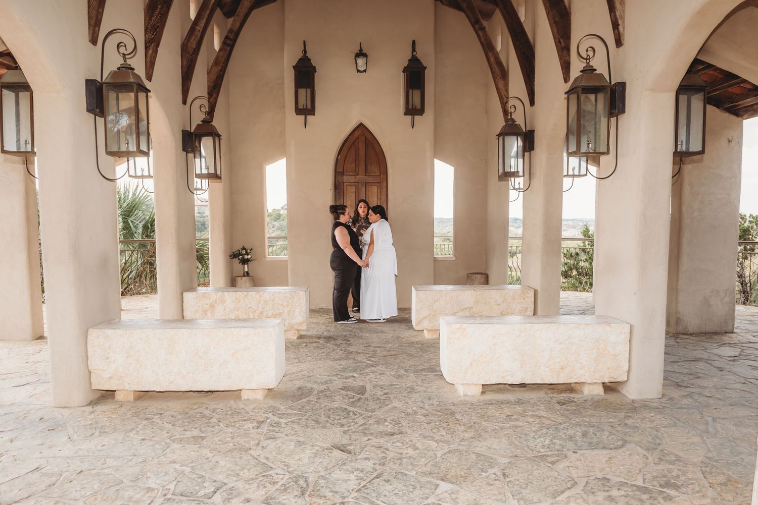 A couple stands intimately framed in stone archway with gothic architecture details and hanging lanterns at a villa venue.