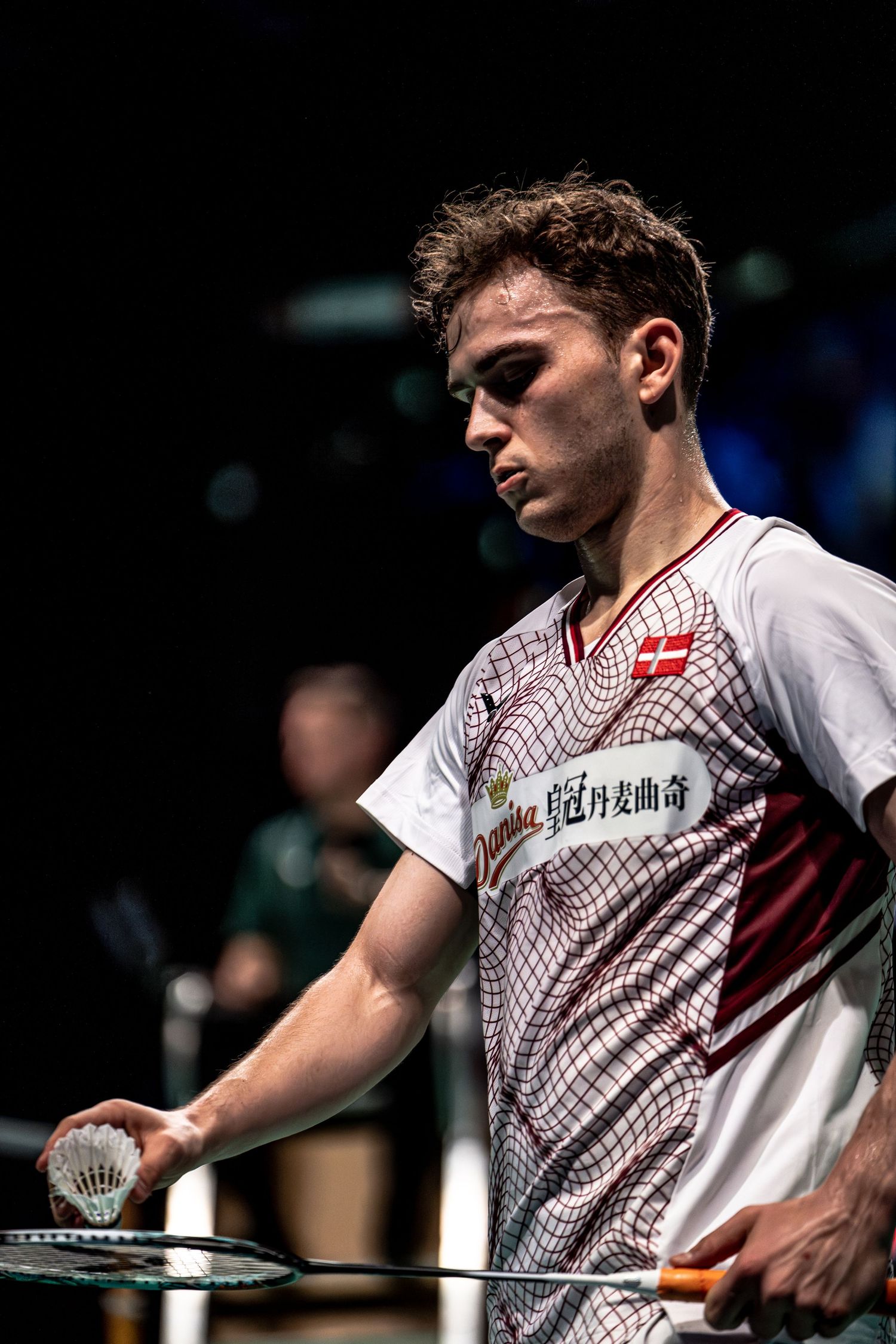 Young male badminton player in Danish team jersey holding shuttlecock and racket during competitive match.
