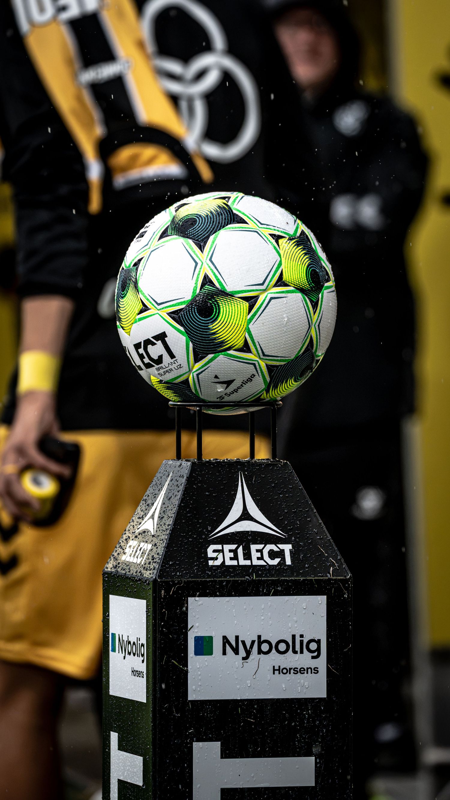 SELECT branded soccer ball on Nybolig Horsens pedestal stand, with players in yellow uniforms visible in background.