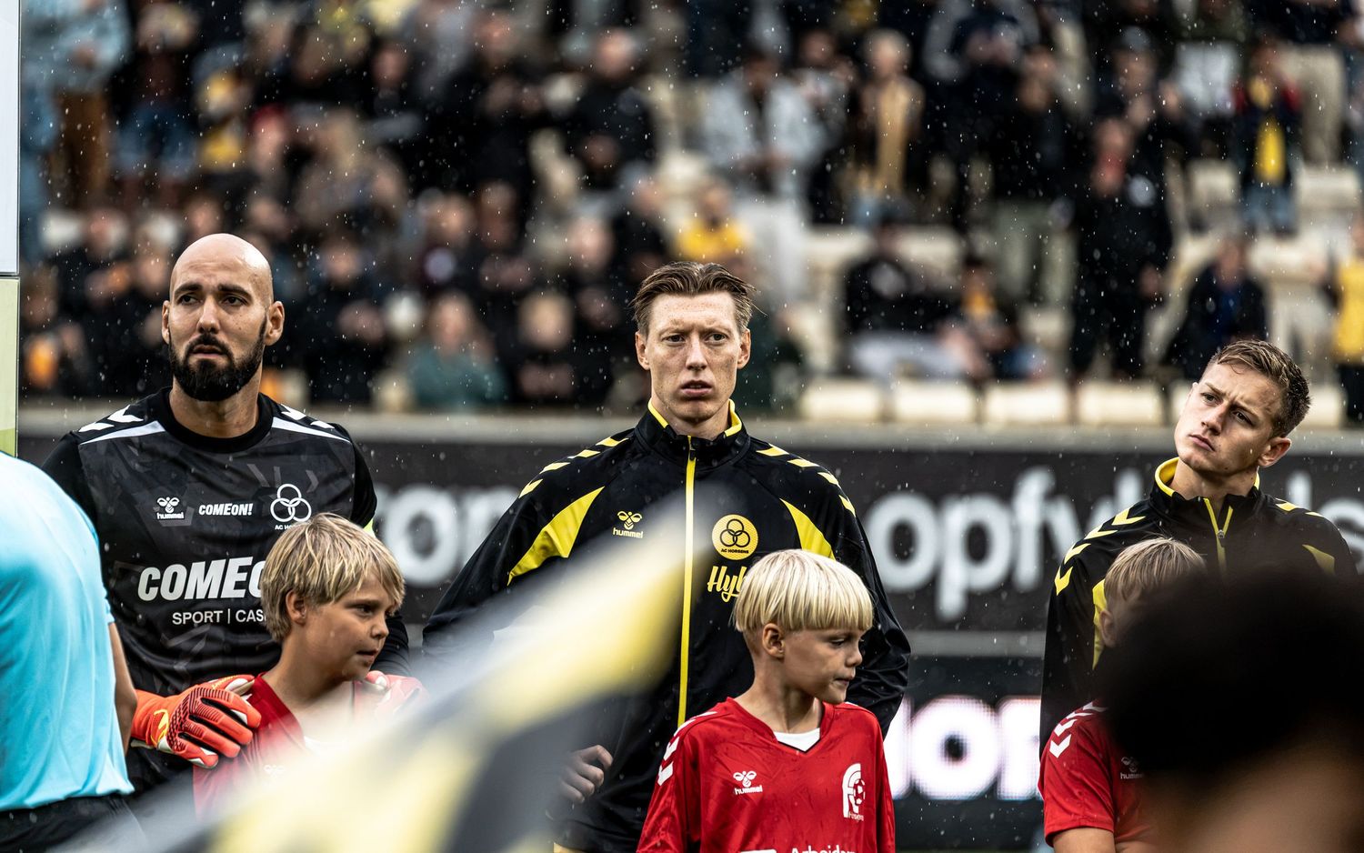 Three football players stand in a line during a match ceremony, wearing black and yellow jerseys with a young mascot beside them.