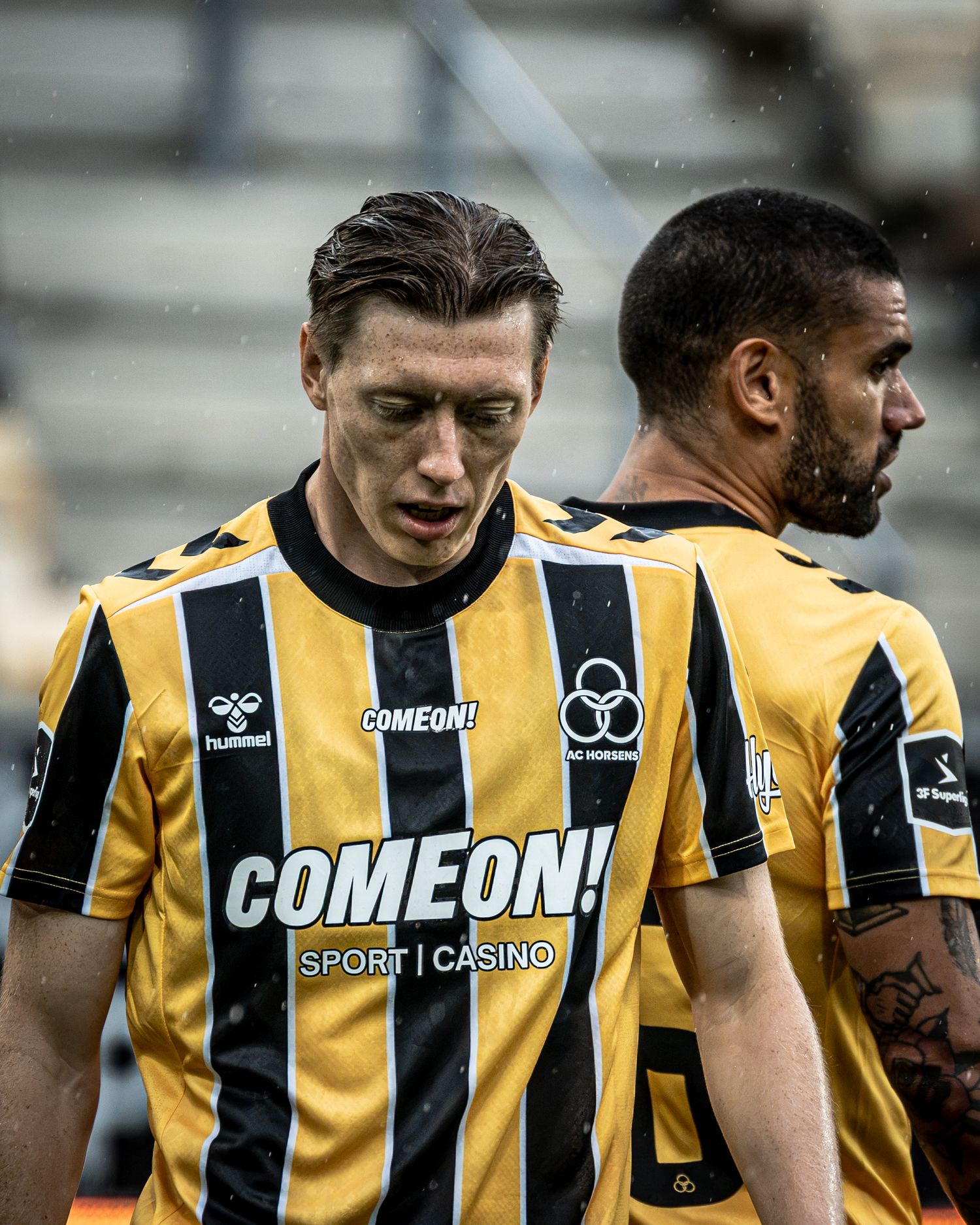 Two soccer players in black and gold Hummel ComeOn! Sport Casino jerseys during a rainy match.