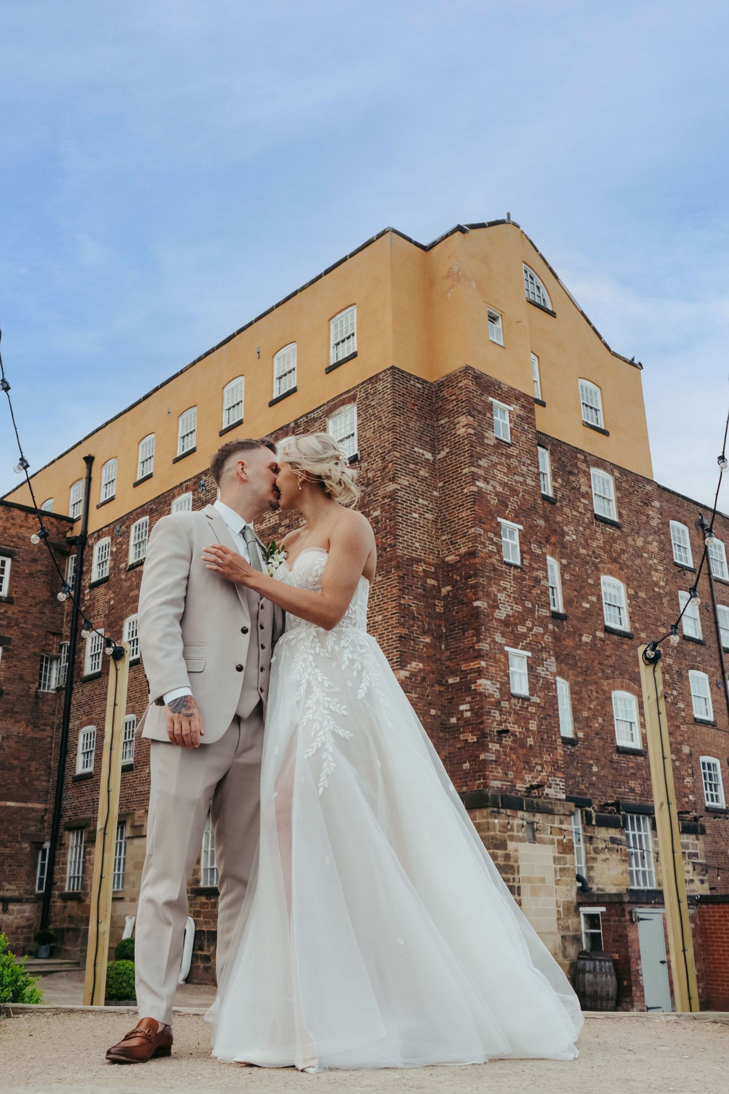 Groom kisses boho style bride as at their wedding reception in industrial derby venue, The West Mill during the summer
