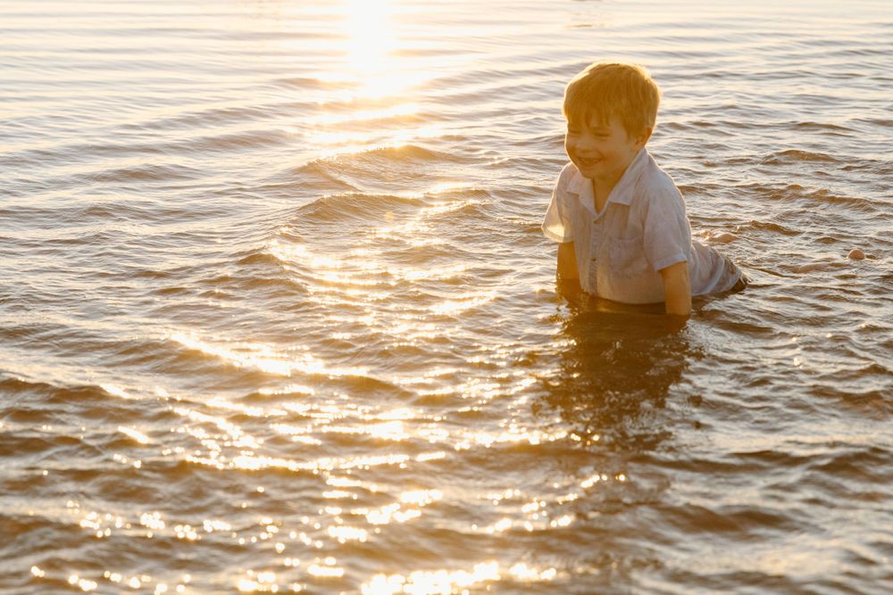 A white-clad figure wades through glittering sunset waters creating ripples across the golden surface.
