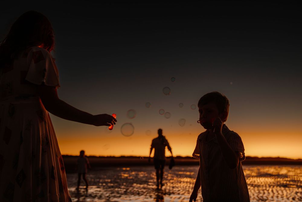 Silhouettes play with bubbles against a dramatic sunset backdrop on a beach.