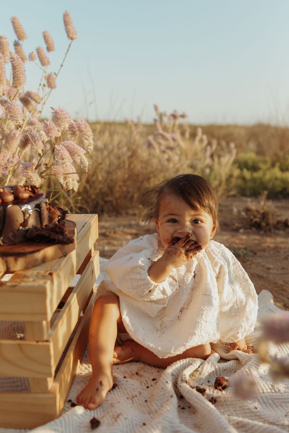 Small child in white dress eating snack outdoors among pampas grass at sunset.