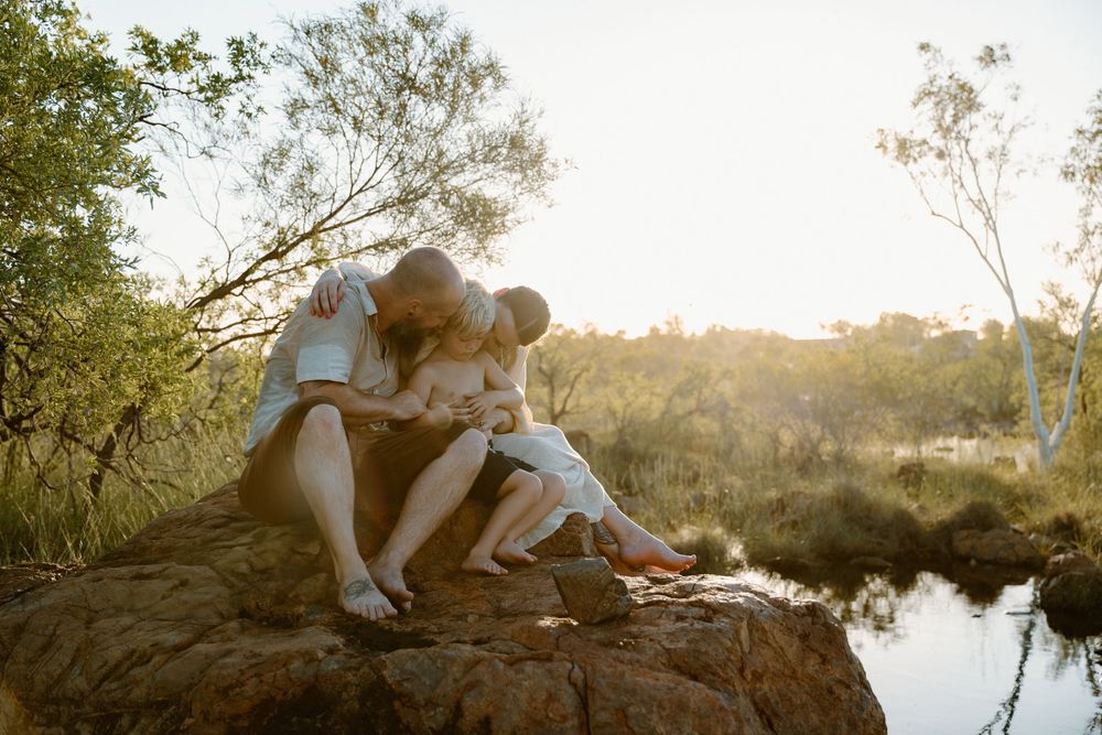 Couple in light summer clothing embraces near tranquil lake at golden hour with trees in background.