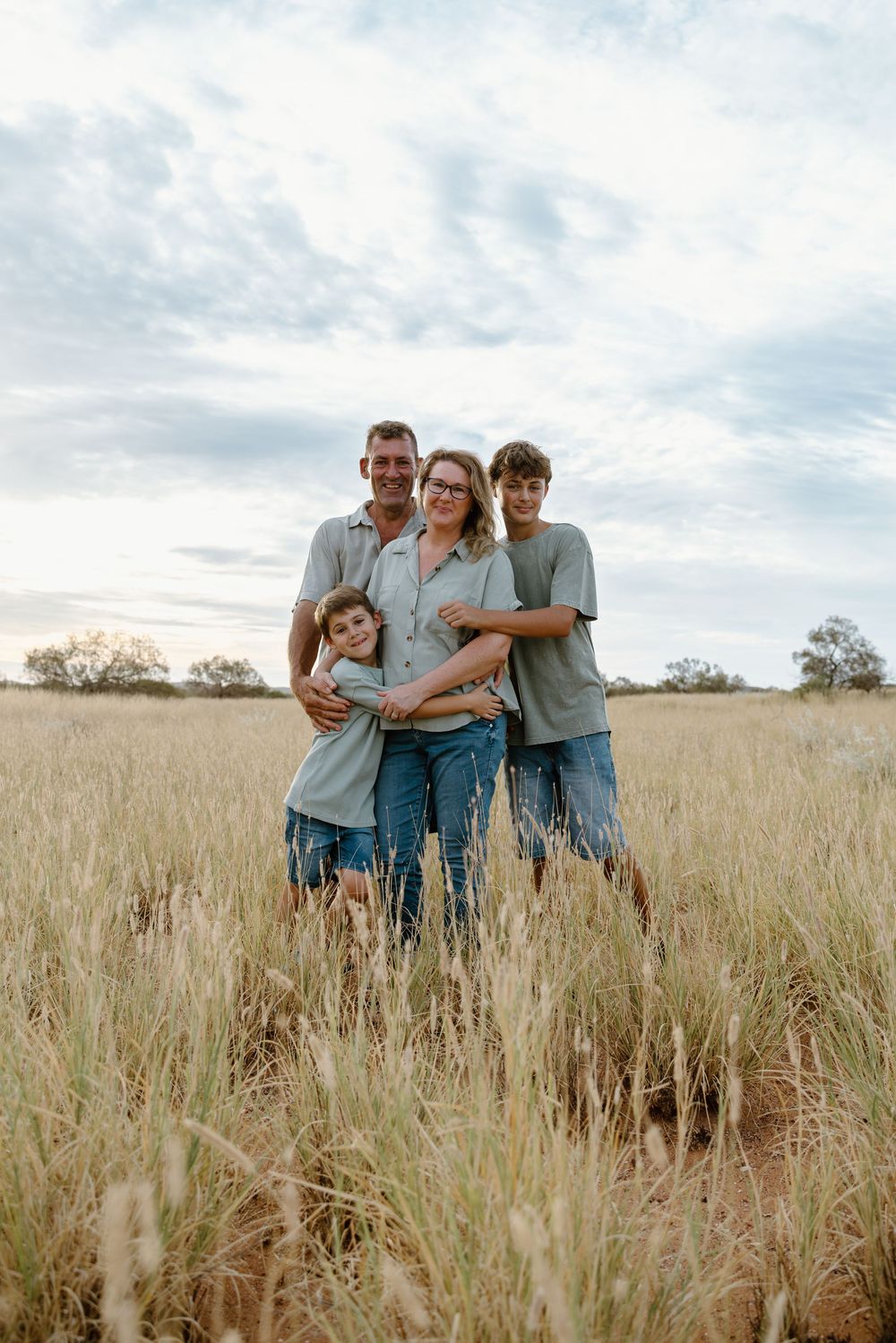 A loving family poses together in a golden field of tall grass during sunset against a cloudy sky.