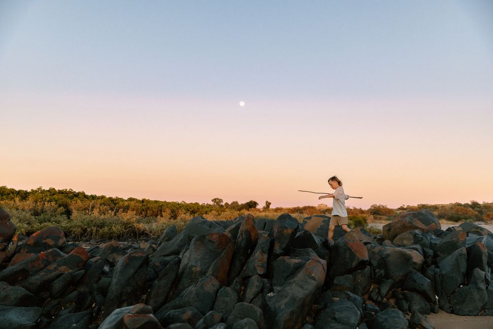 A silhouetted figure stands on rocky terrain against a sunset sky with a moon visible in the distance.
