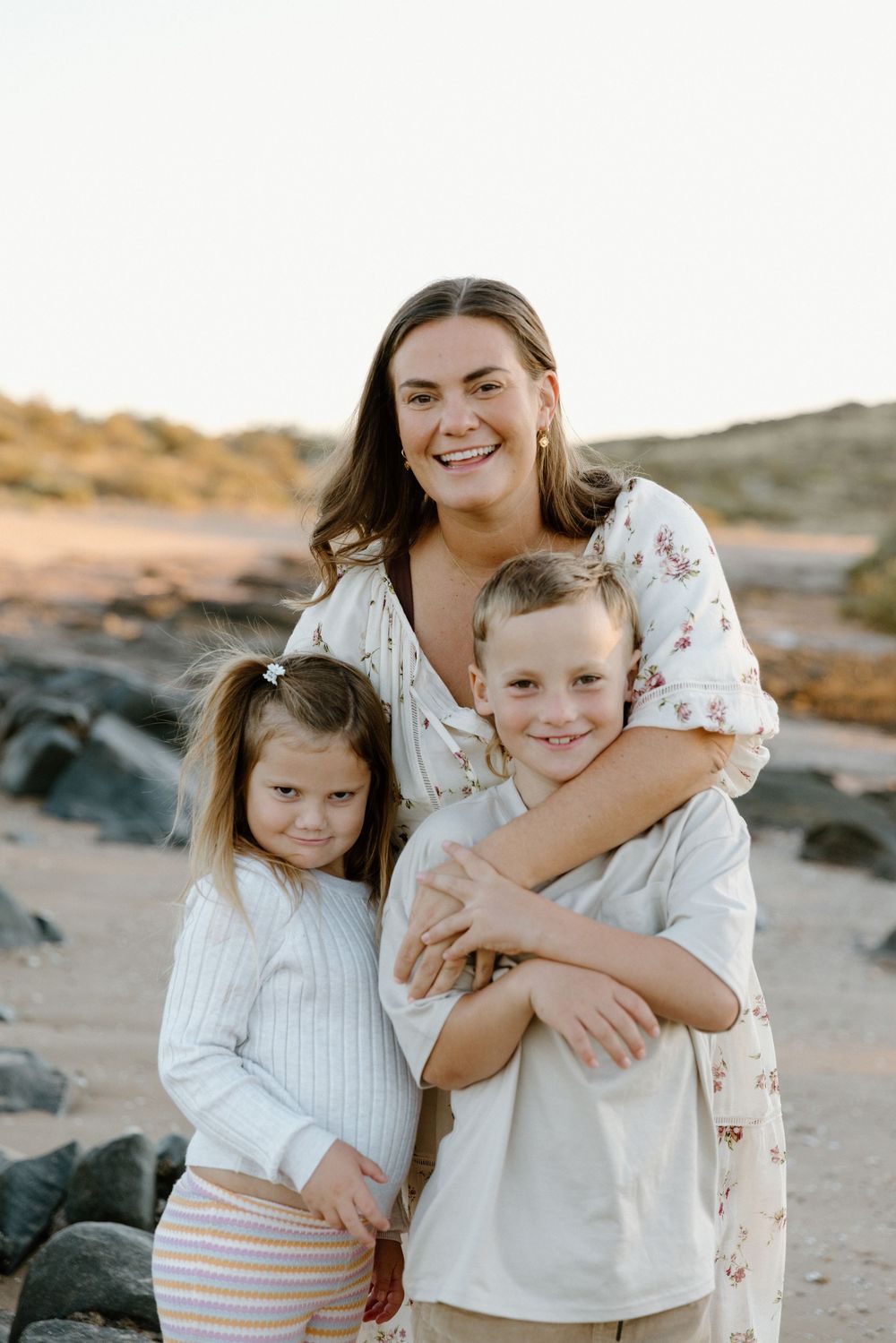A loving family portrait of a woman with two children posing together on a rocky beach at sunset.