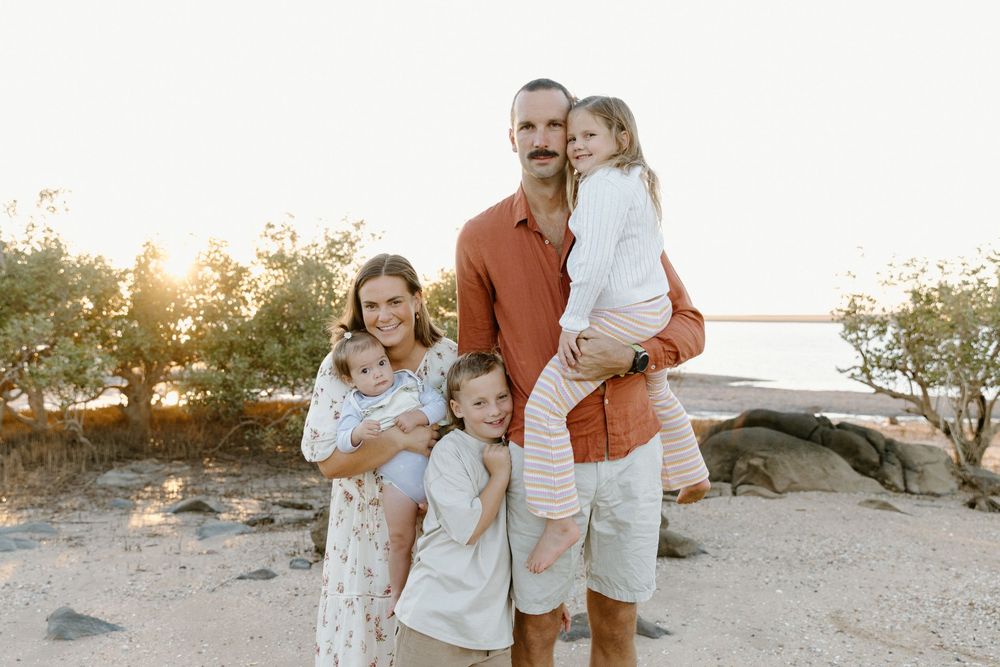 A family poses together on a sandy beach at sunset with trees and water in the background.