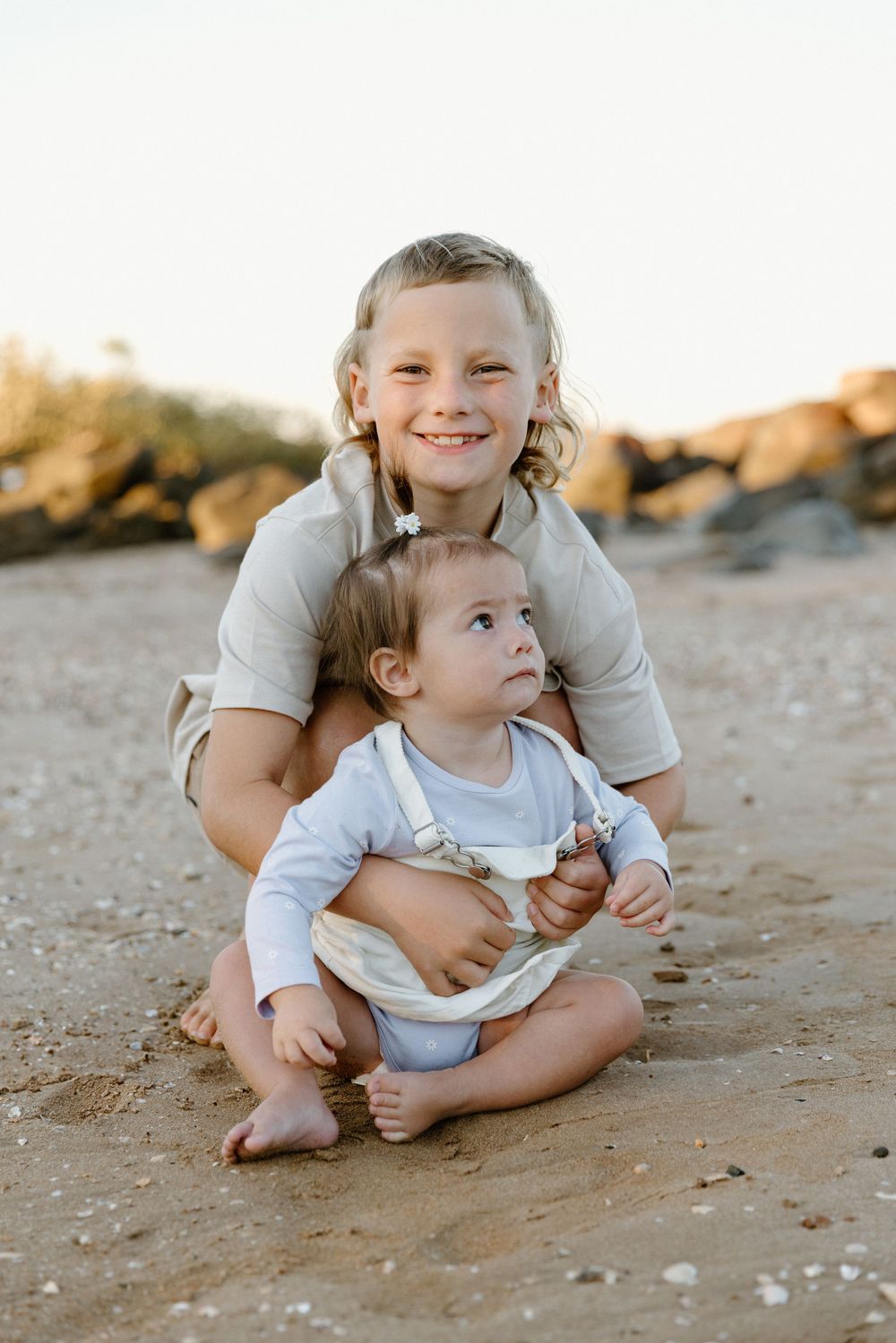 Two children sitting together on a sandy beach at sunset with rocks in the background.