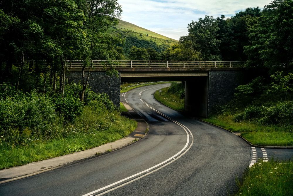 Winding road passes under stone bridge through lush green countryside hills.
