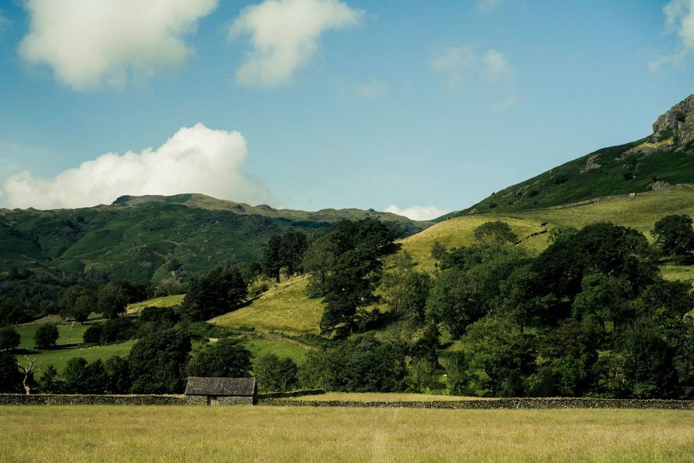 Rolling green hills and mountains under blue sky with scattered clouds in countryside landscape.