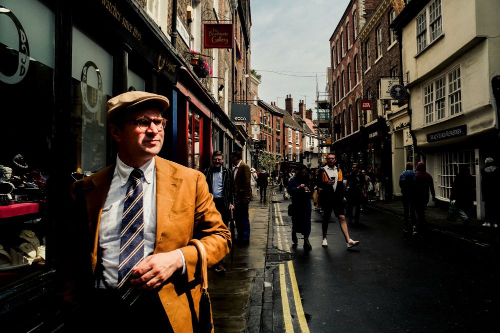 People walking down a narrow historic street with brick buildings and shops in England.