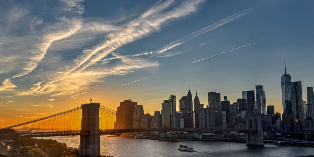 Manhattan skyline and Brooklyn Bridge silhouetted against dramatic sunset sky.
