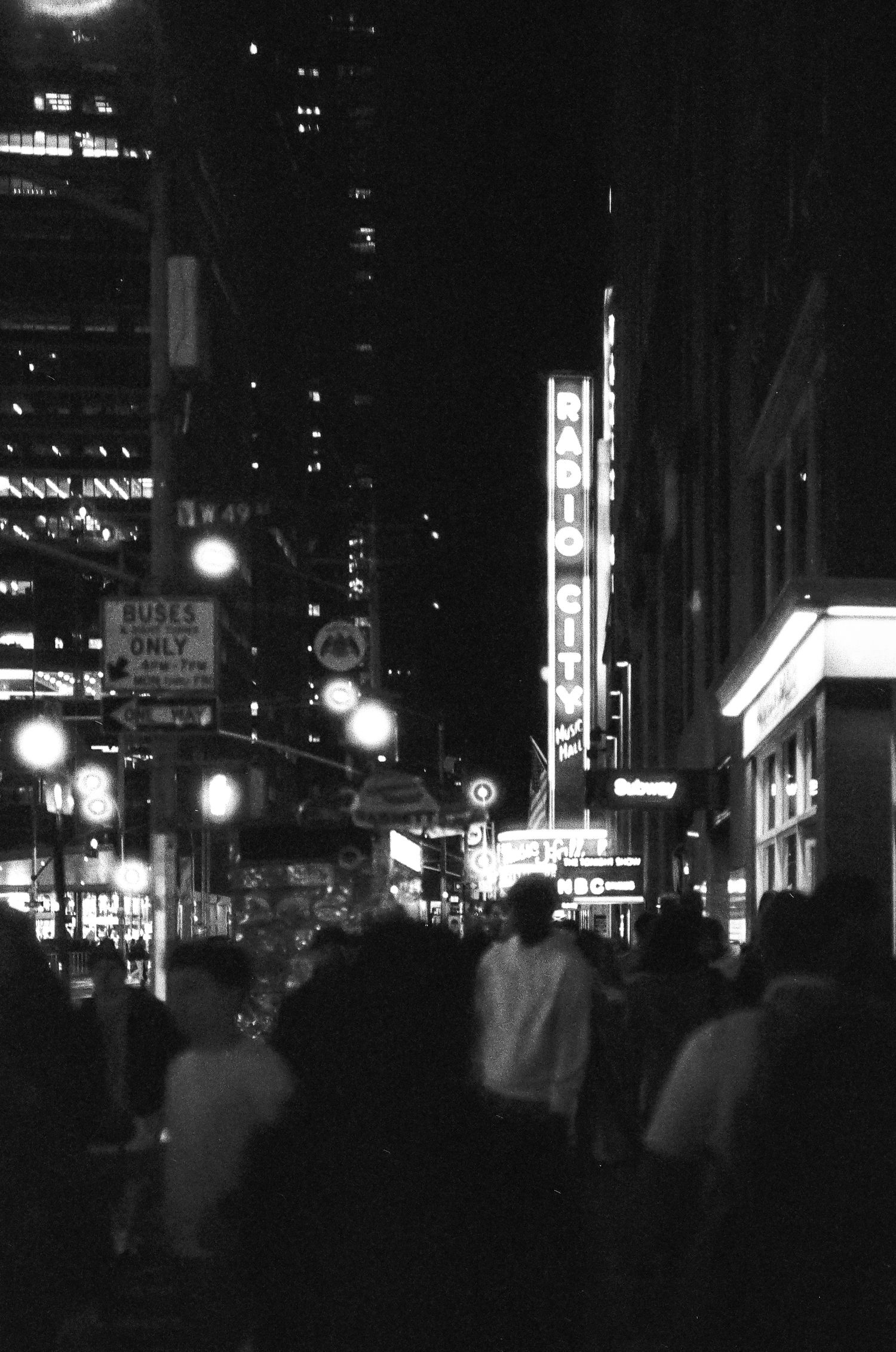 Moody black and white cityscape of illuminated signs and crowds on a busy night street.