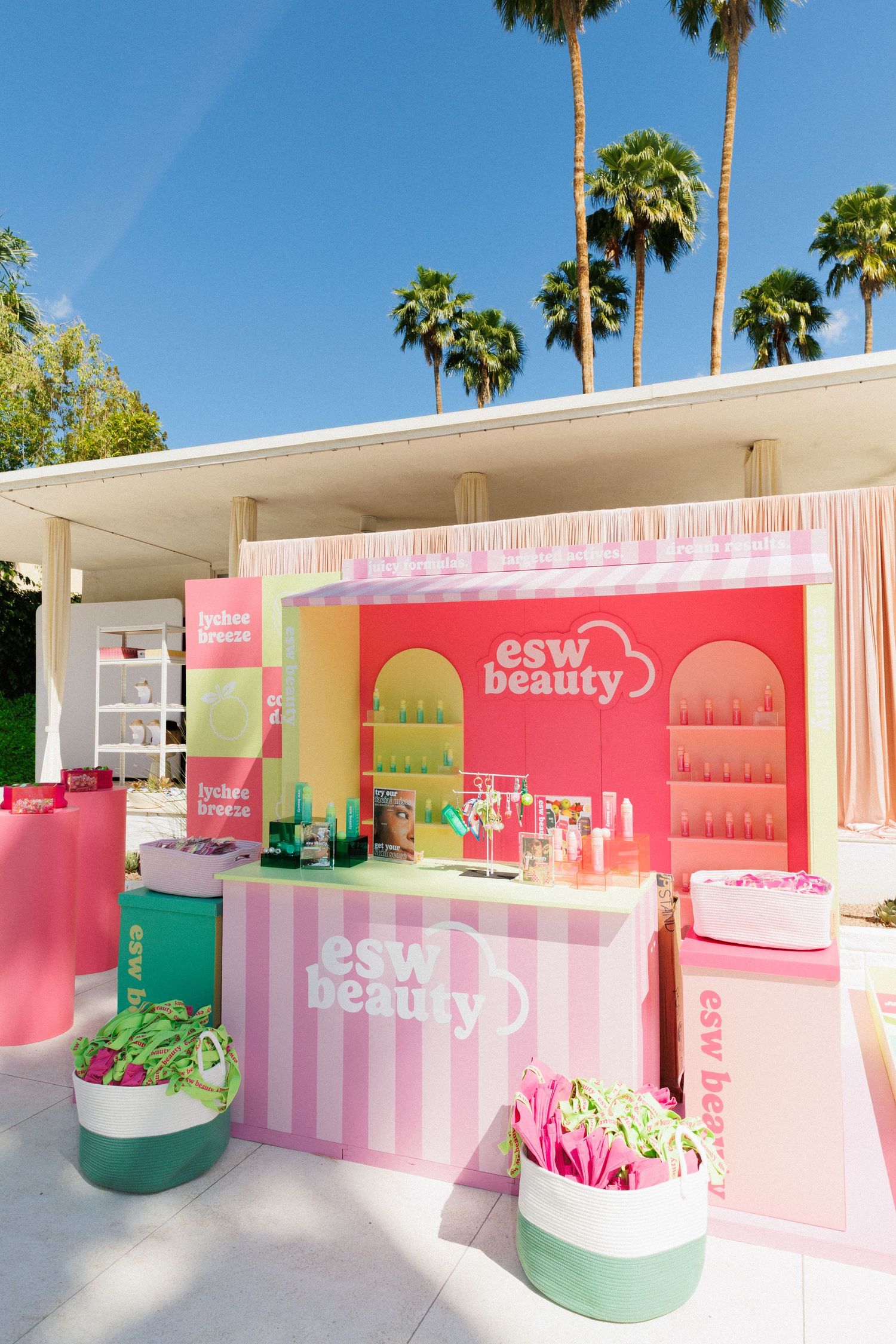 Colorful ESW Beauty outdoor pop-up booth with pink striped design, product displays, and palm trees at a sunny event.