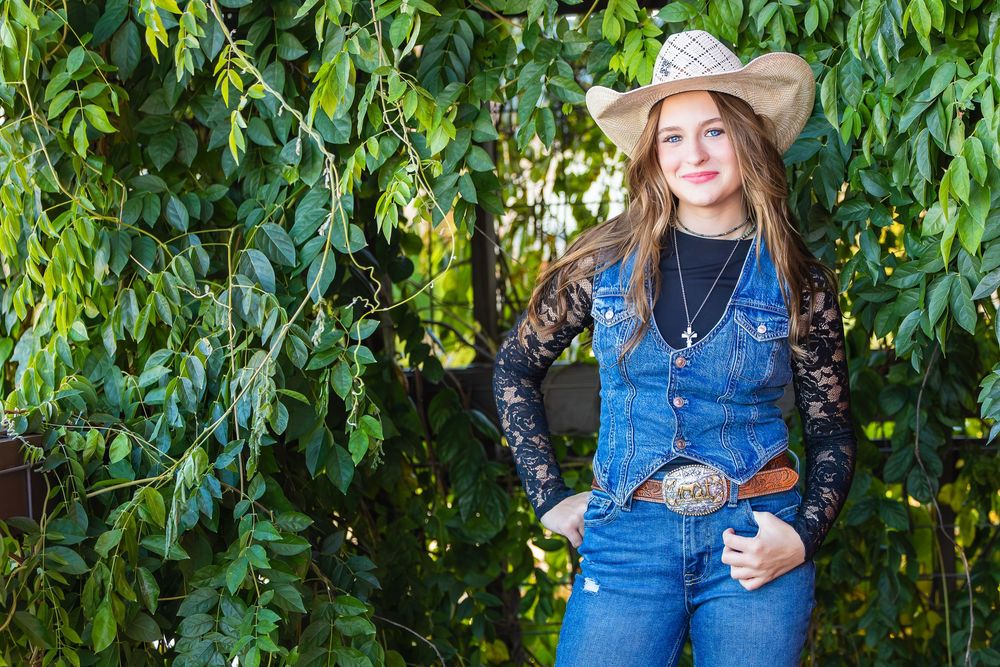 Country western style with denim overalls, black lace sleeves, and cowboy hat posed against green foliage backdrop.