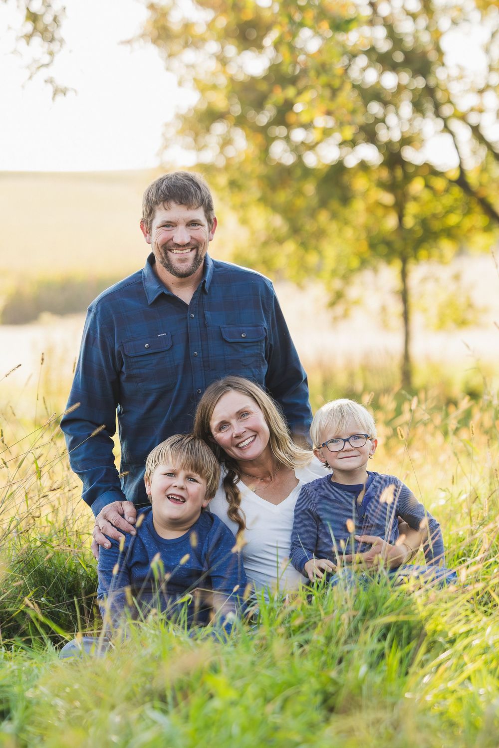 A family sits together in tall grass during golden hour with sunlit trees in the background.