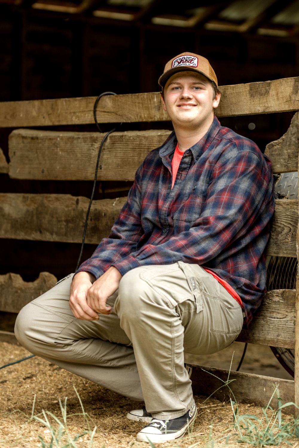 Casual portrait in plaid flannel and baseball cap sitting on wooden structure.