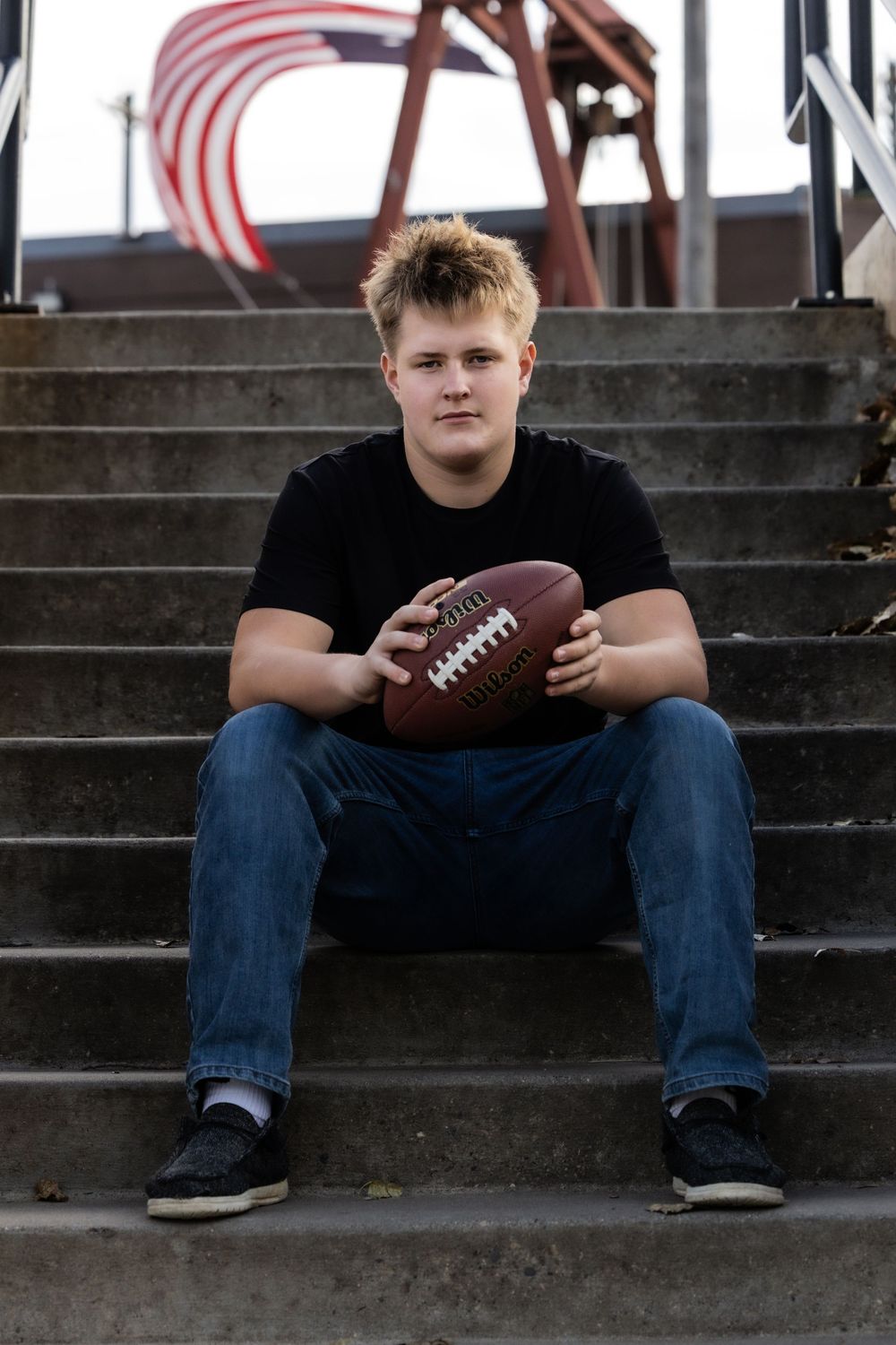 A young football player sits on concrete stadium steps holding a football while wearing jeans and a black shirt.