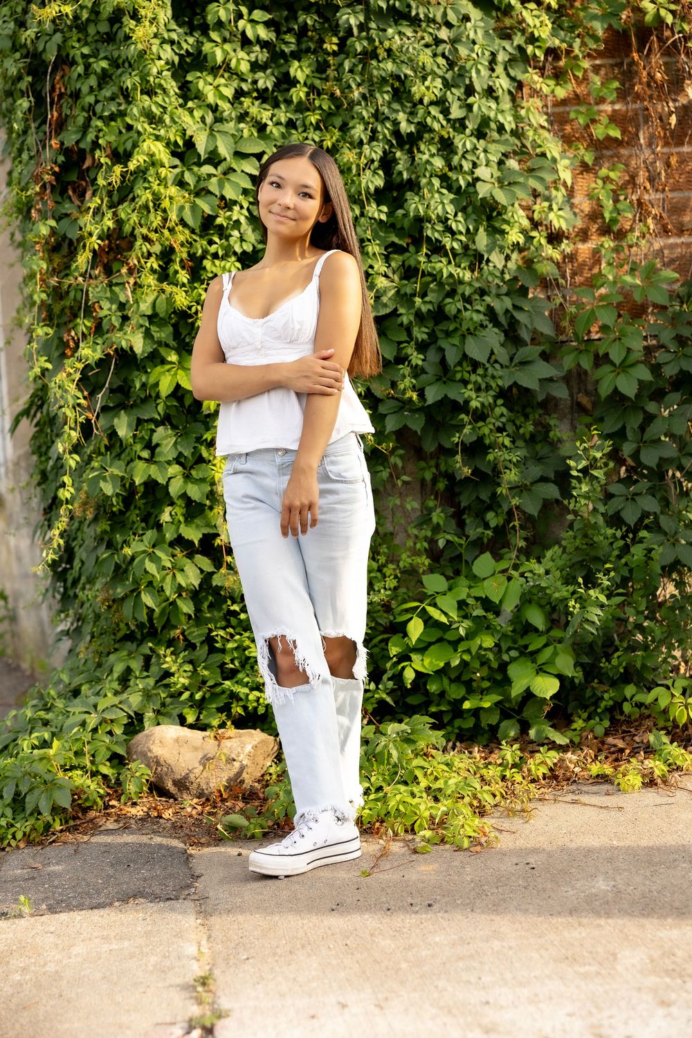 Person in white summer outfit and sneakers poses casually against lush green vine-covered wall outdoors.