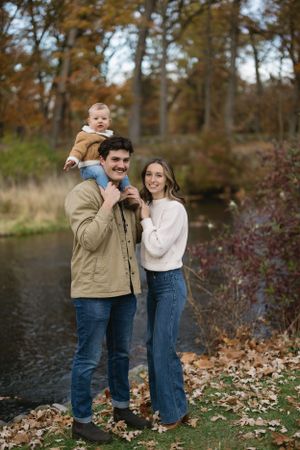 A family of three poses together near an autumn pond with fallen leaves.