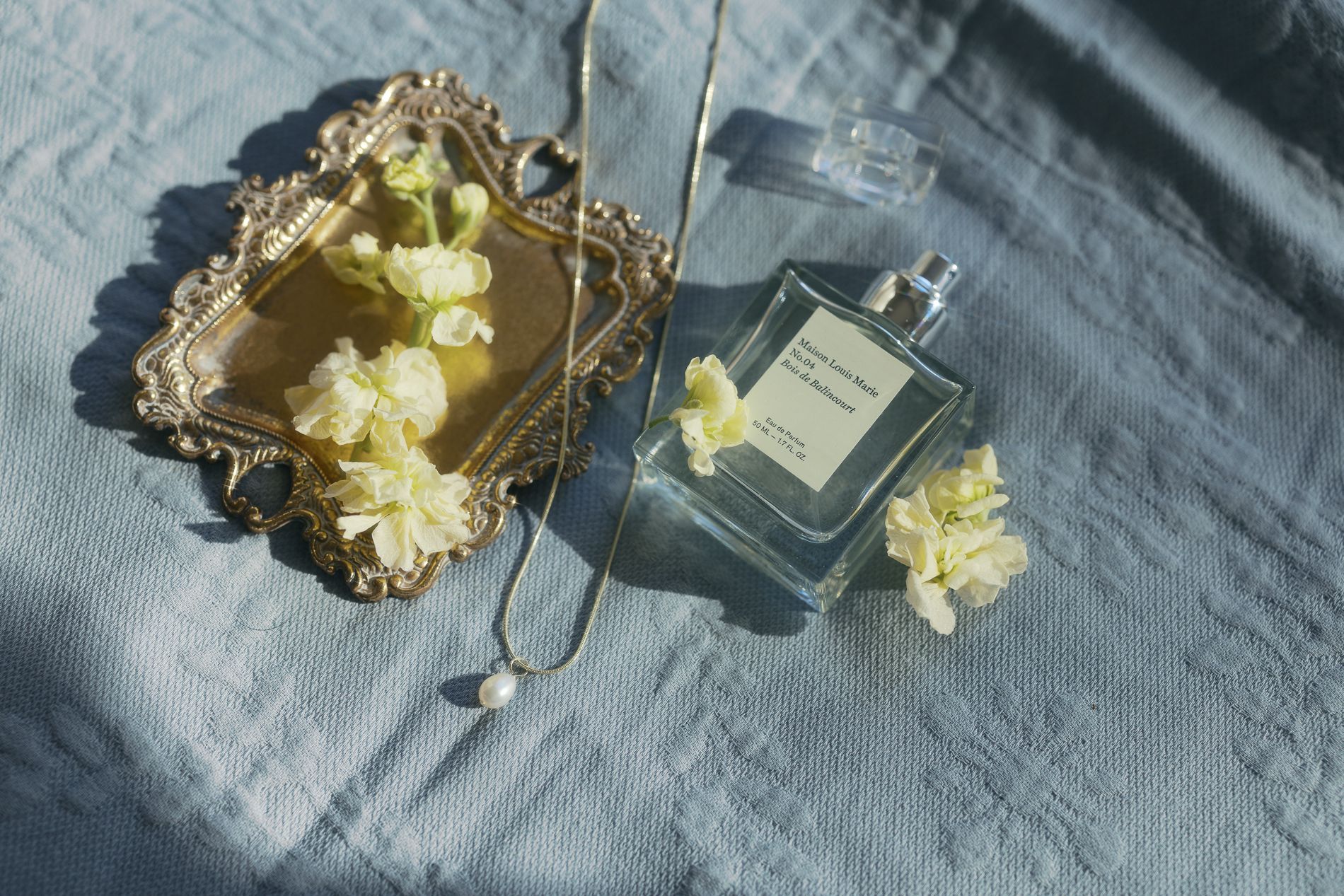 Flatlay composition of perfume bottles, white flowers, and jewelry arranged on gray textured fabric.