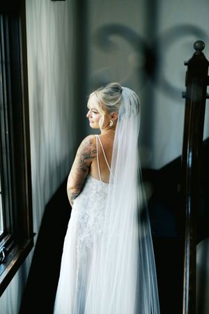 A bride in a white lace wedding gown with a flowing veil stands by a window creating dramatic shadows on the wall.