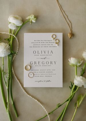 Elegant wedding invitation with white ranunculus flowers, pearl necklace and gold rings on beige background.